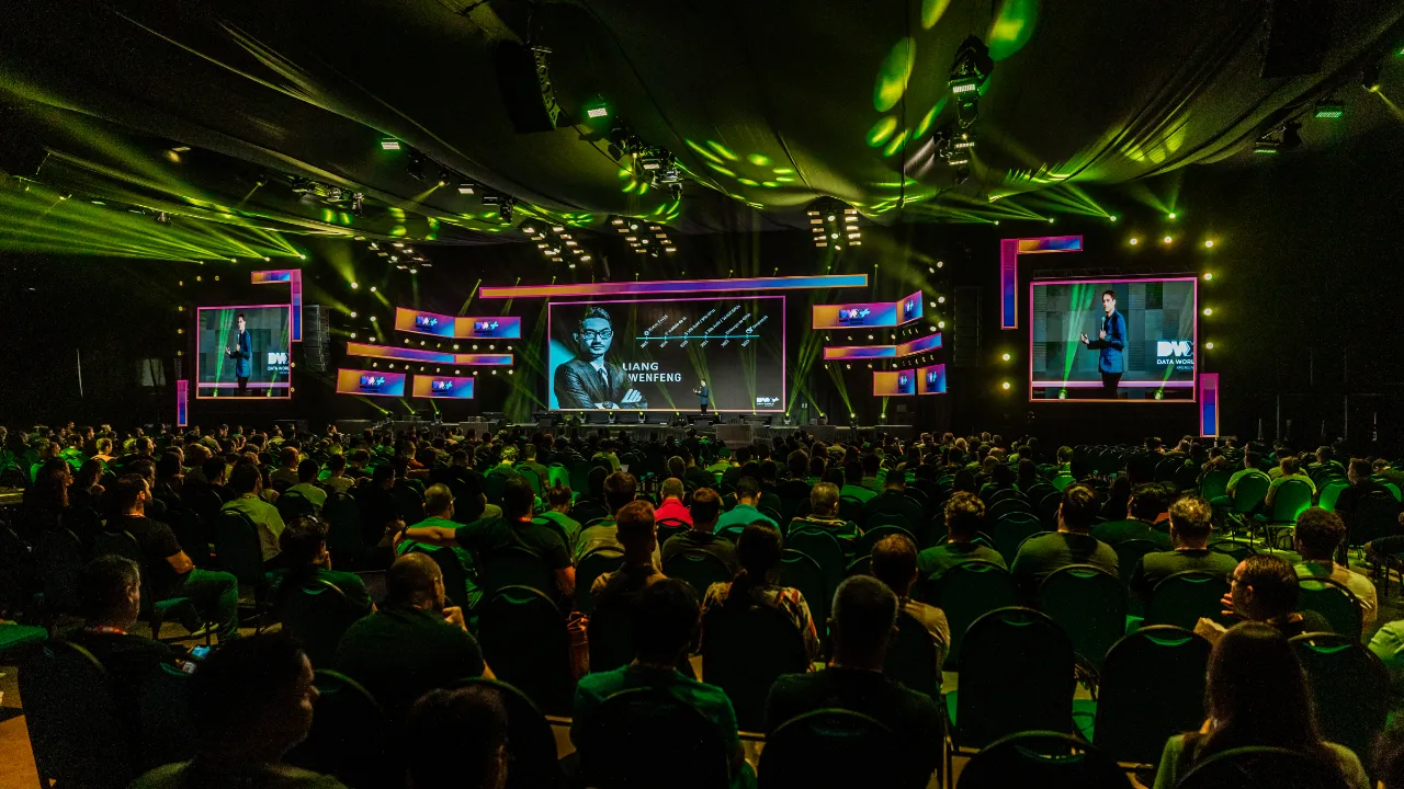 Magno Maciel palestrando em um grande auditório lotado durante o evento Data World Xperience 25, com iluminação cênica e telões exibindo sua apresentação.